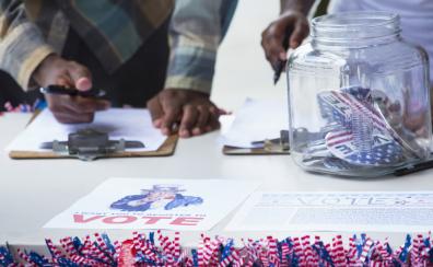 Two people stand behind a table with a VOTE sign on it. They are writing on a clipboard. 