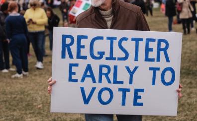 A man holds a hand-painted sign that reads "Register early to vote"