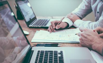 Two white men sit at a desk, with Mac laptops open in front of them. They are conferring over, and look to be editing, a stack of papers in between them.